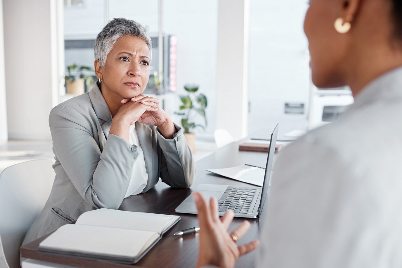 Woman with hands folded under her chin sits behind a desk with open notebook and laptop looking concerned while another person who is only partially visible speaks to her and gesticulates 