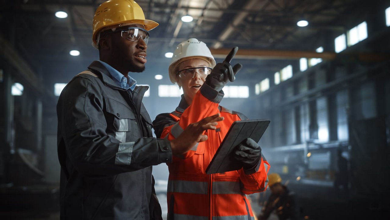 Two workers in hard hats looking at a tablet in a factory.