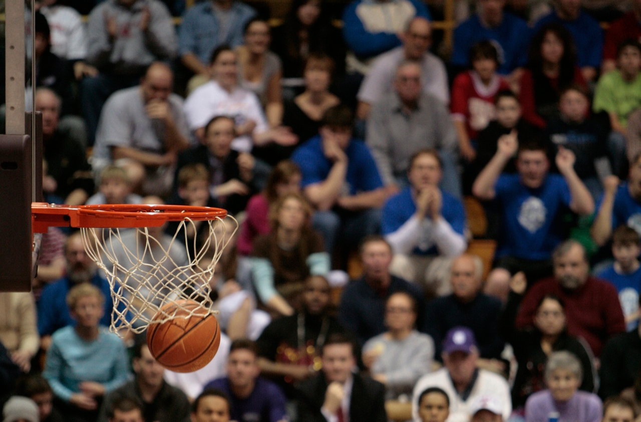 Basketball fans sitting on the blechers show emotion as a basketball falls through the basket and net to score.