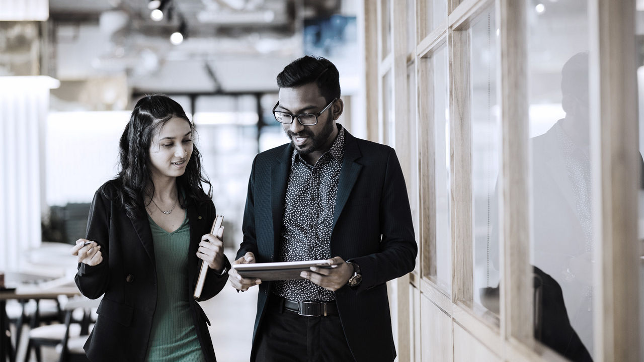 A man in a suit looks at a tablet in an office, as he walks along side a woman in a blazer and they discuss what he's looking at on the tablet.