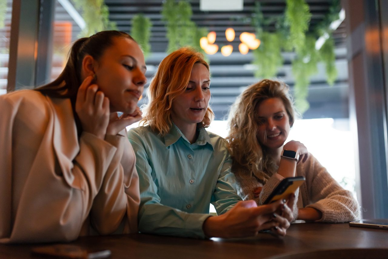 Three businesswomen share ideas and enjoy each other's company in a stylish cafe, focused on their phones as they collaborate on projects in a vibrant atmosphere.
