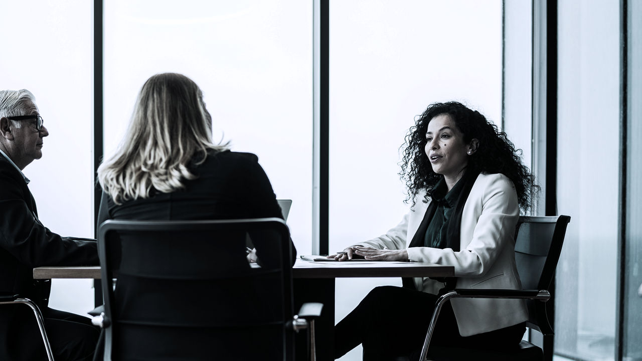 Female leader sitting at table on right speaking to two colleagues