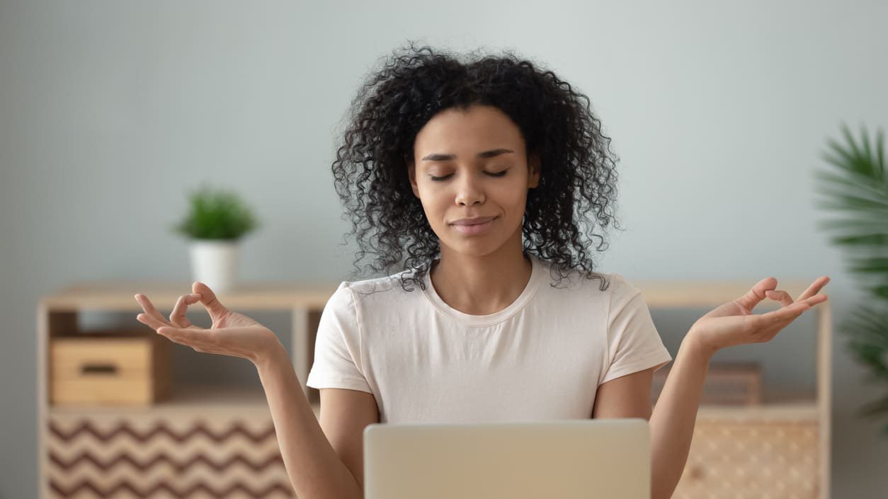 A woman meditating in front of her laptop at home.