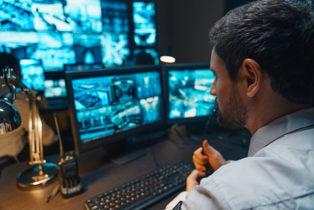 Security  Controller with Headset Talk on a Call in security office. Office Room is Full of Desktop Computer Displays with Navigation Screens, Data for the Team.