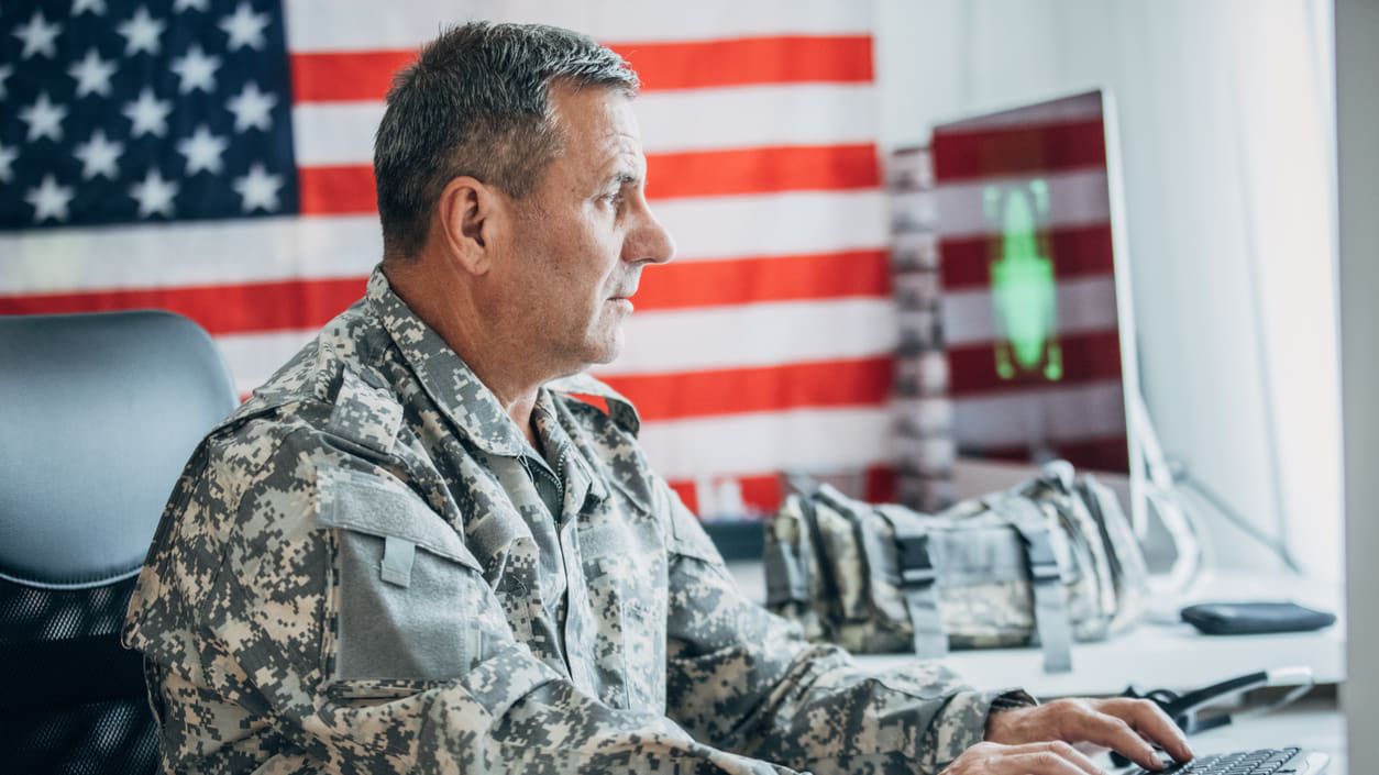 A man in military uniform using a computer in front of an american flag.