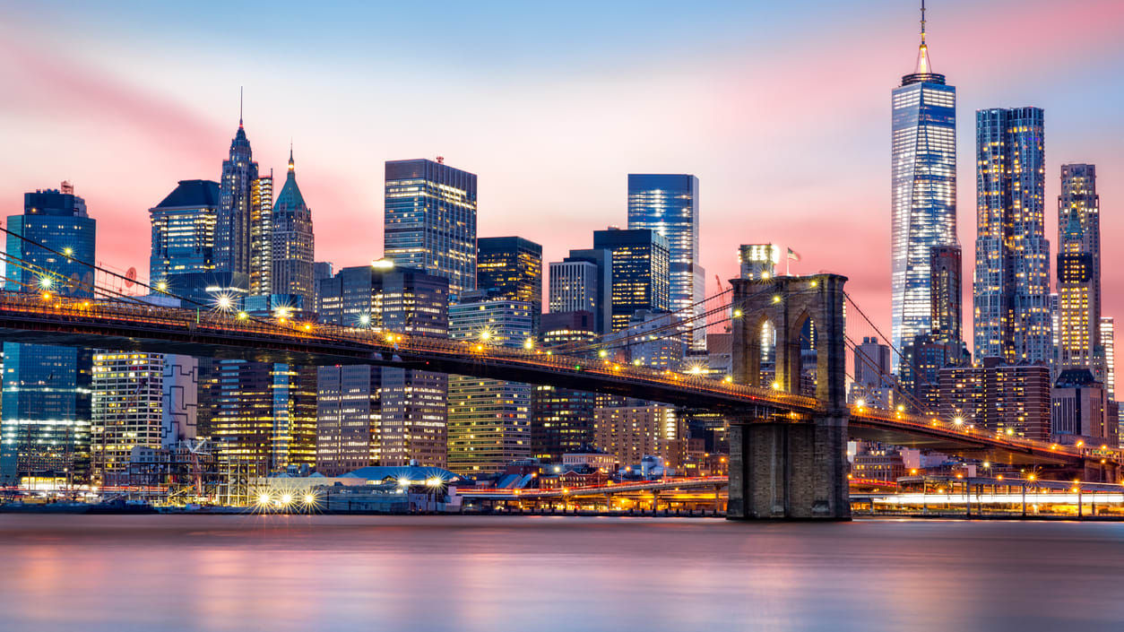 New York City skyline at sunset with the Brooklyn Bridge in the foreground and illuminated skyscrapers reflected on the East River.