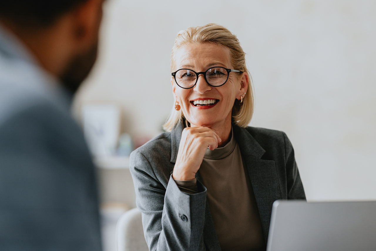 Smiling senior businesswoman in glasses conversing with a colleague during an office meeting. Professional and positive work environment.