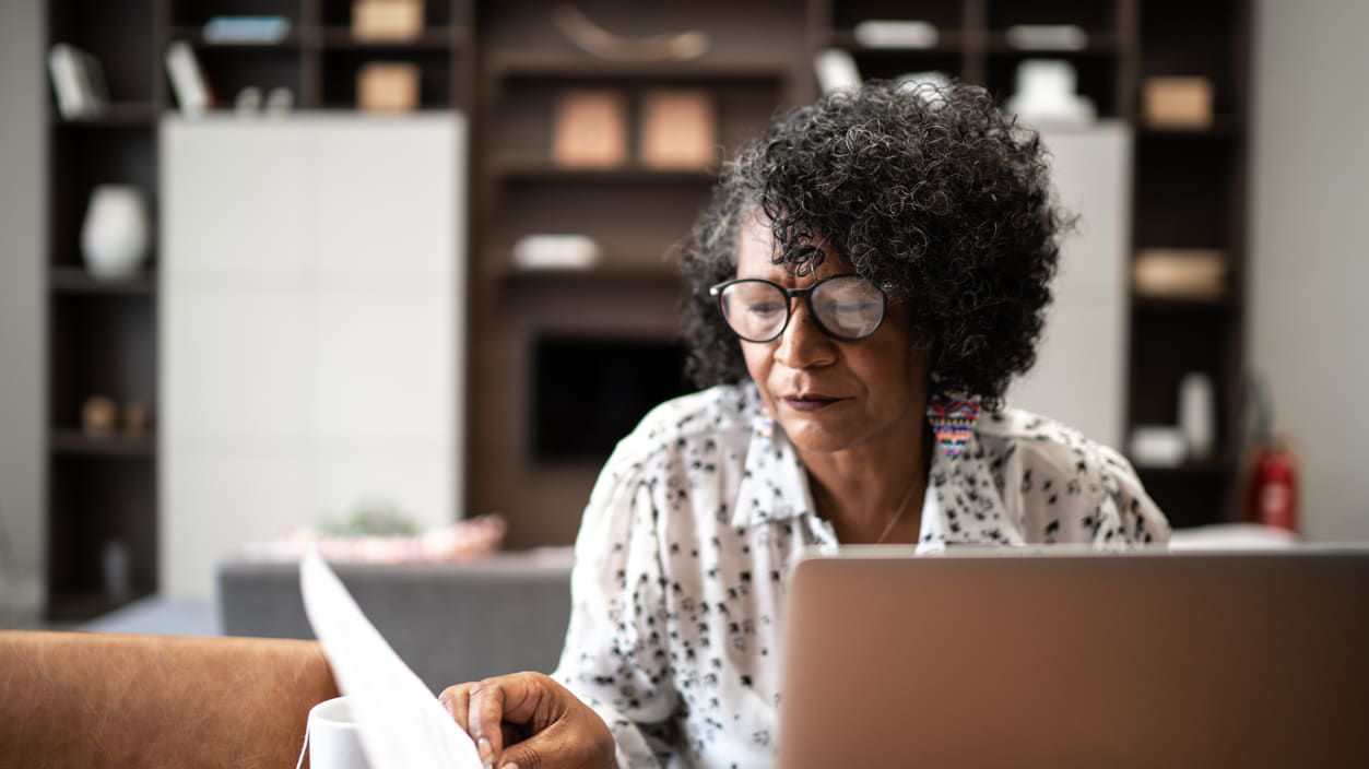 An older worker at her desk leafing through paperwork behind her computer, looking through her glasses