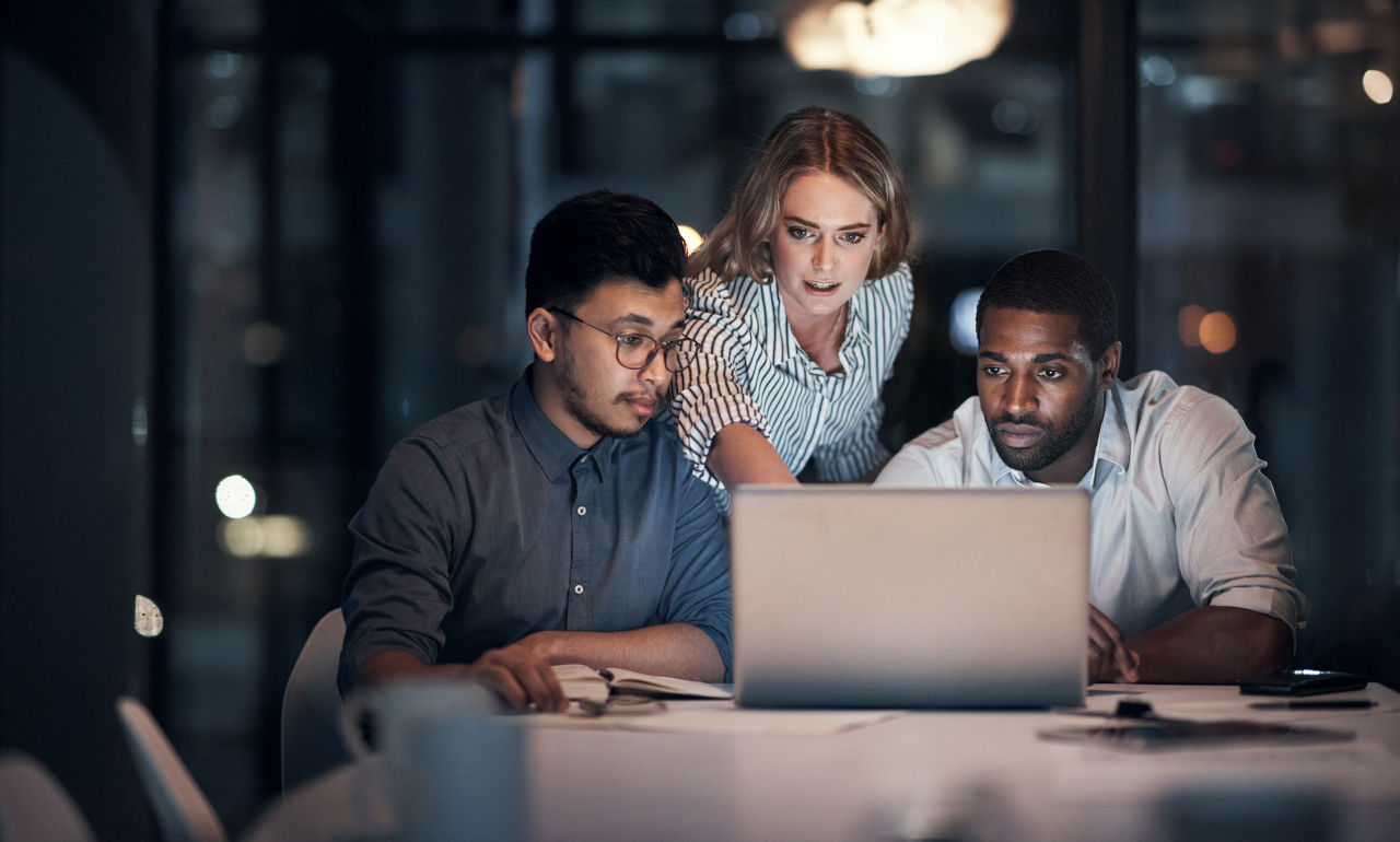 A female employee points at a shared computer as she and two male colleagues work overtime.