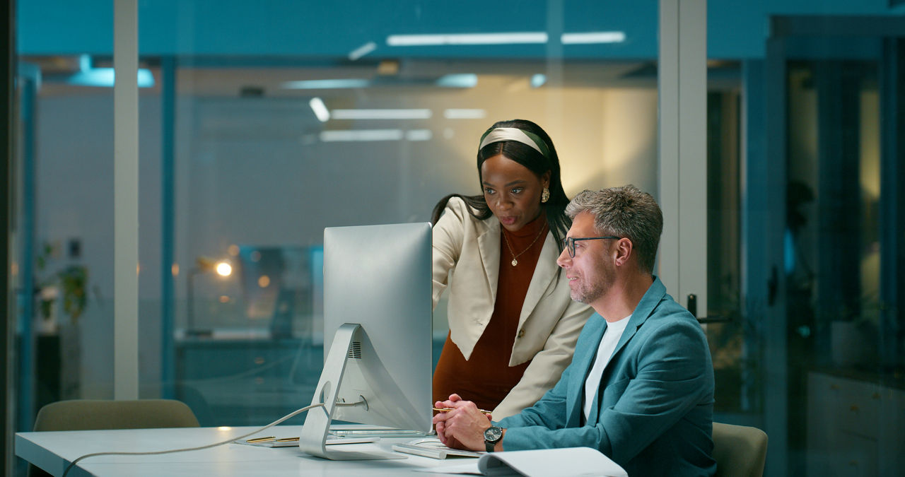 Colleagues, pointing at a computer, go over company strategy at night. Financial analyst, man and woman with tech, working late and planning for funding opportunity.