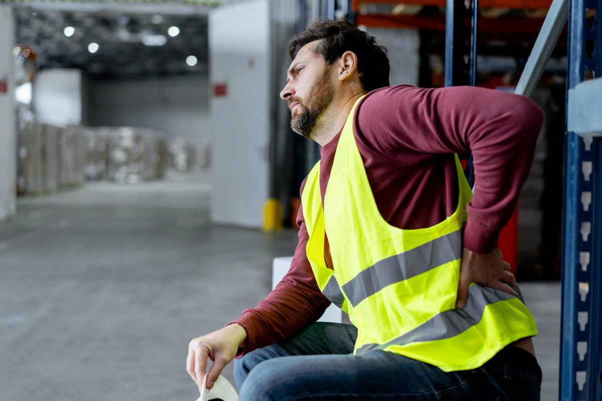Upset, tired, man, pain foreman wearing workwear touching back, having pain, spasm, sitting in warehouse after work. Concept of treatment, health care