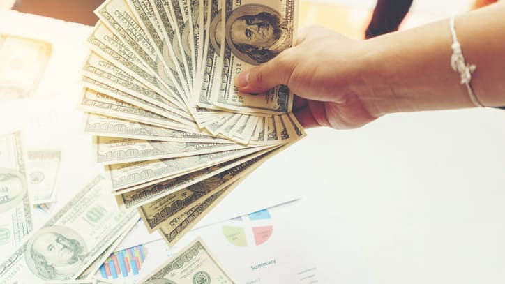 A hand is holding a stack of money in front of a desk.