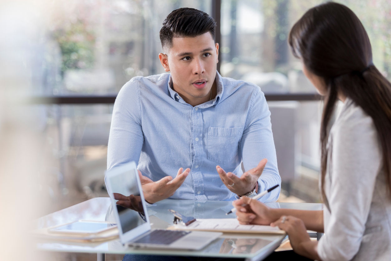 A young woman and man talk over a table, holding a computer, tablet, and paperwork. The man gestures with his hands, as the female listens.