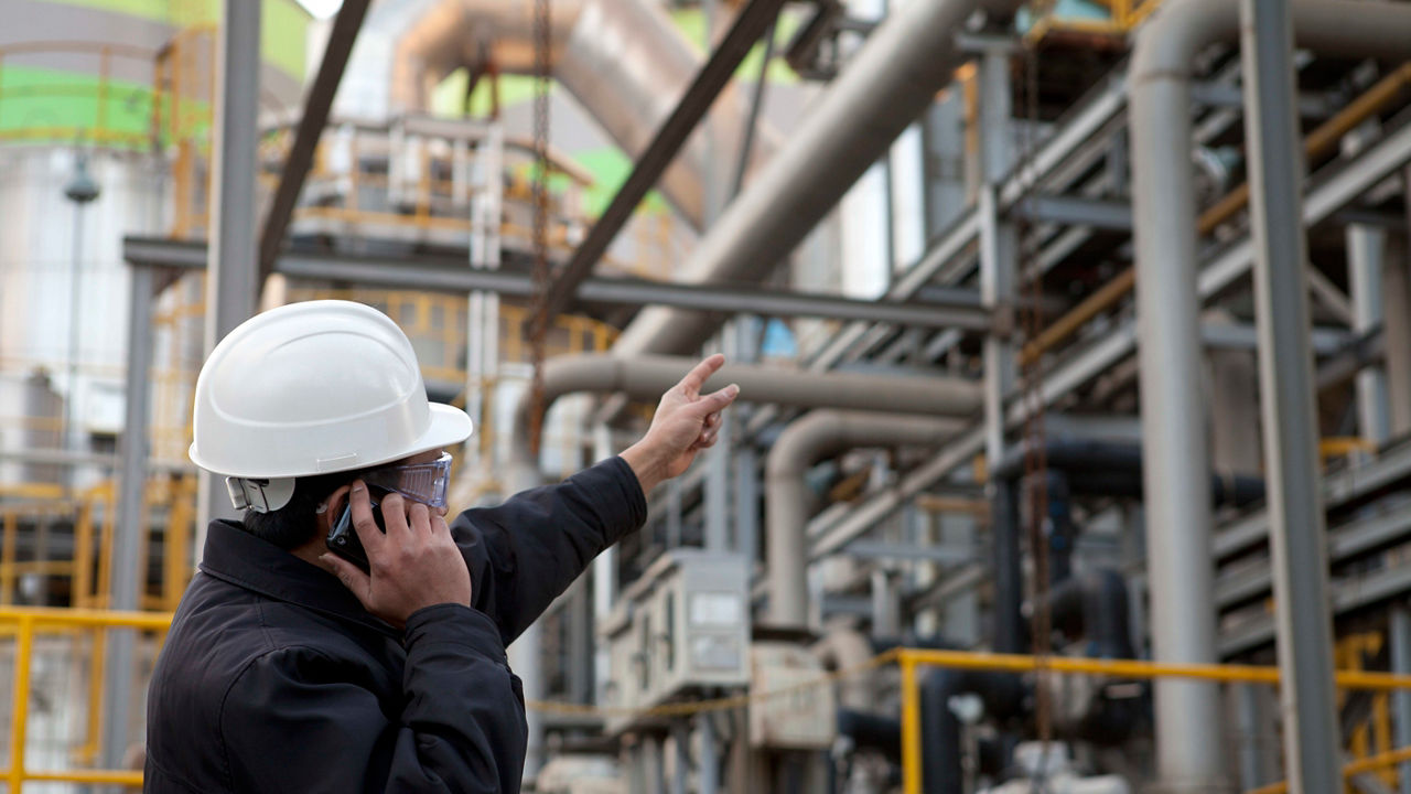 A man in a hard hat is talking on the phone at an oil refinery.