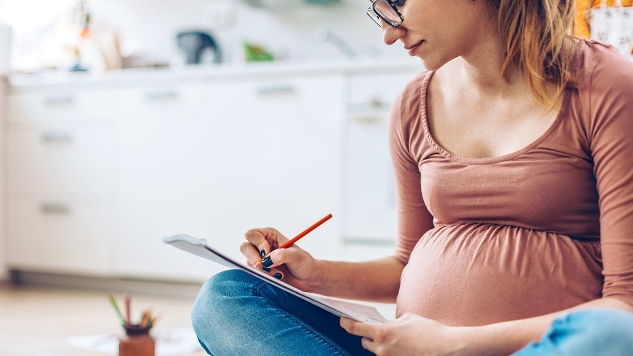 A pregnant woman sits on the floor and writes on a clipboard.