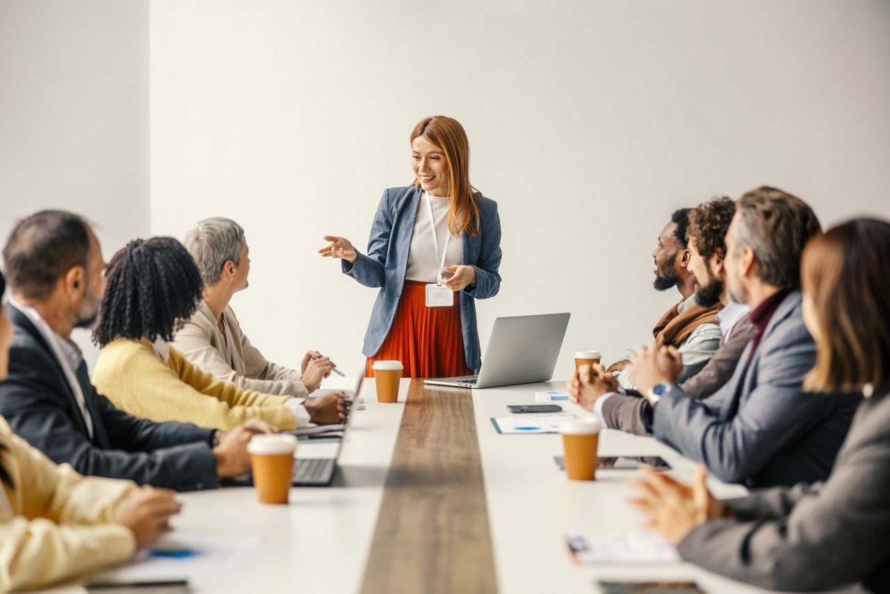Young woman presenting to a diverse business team in a modern office, colleagues listening attentively during a corporate meeting focused on strategy, collaboration and growth