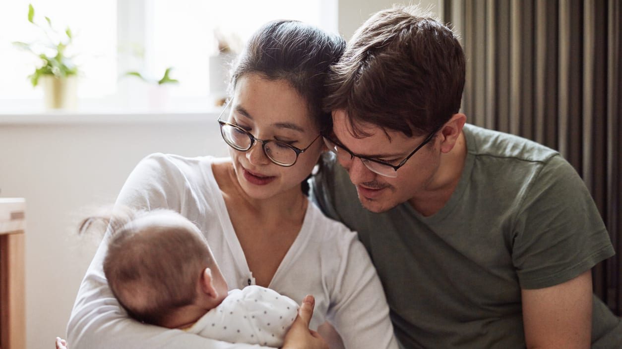 A mother holding her infant with the father looking over the mother's left shoulder at the baby