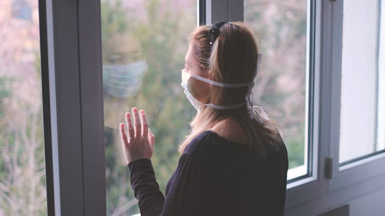 A woman wearing a surgical mask looking out of a window.