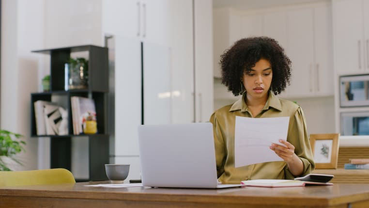 Woman sitting at a table with a laptop, reviewing a document with a concerned expression in a bright home office or kitchen workspace.