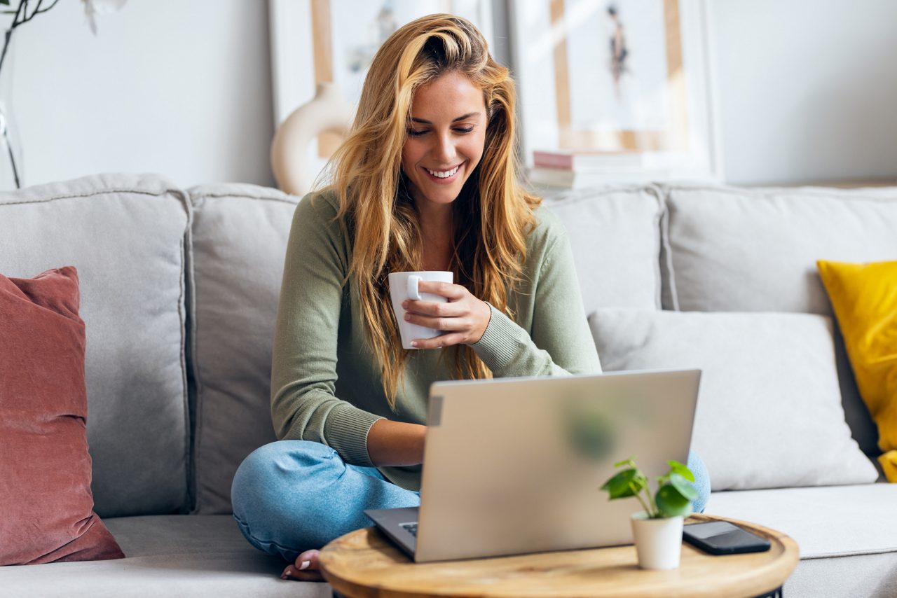 Shot of beautiful young woman working with her laptop while drinking a cup of coffee sitting on a couch at home