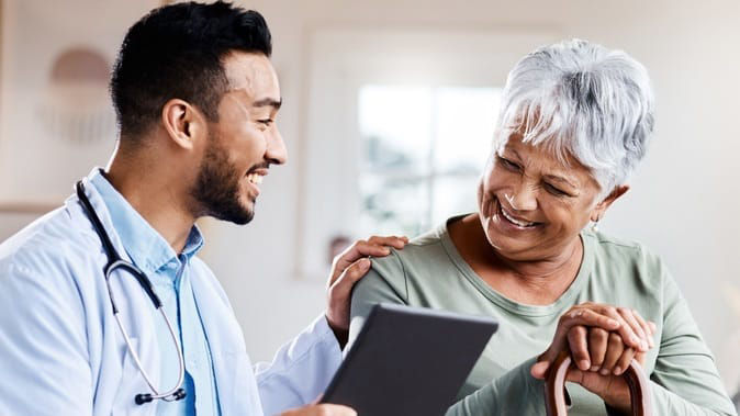 A doctor is talking to an elderly woman while holding a tablet.