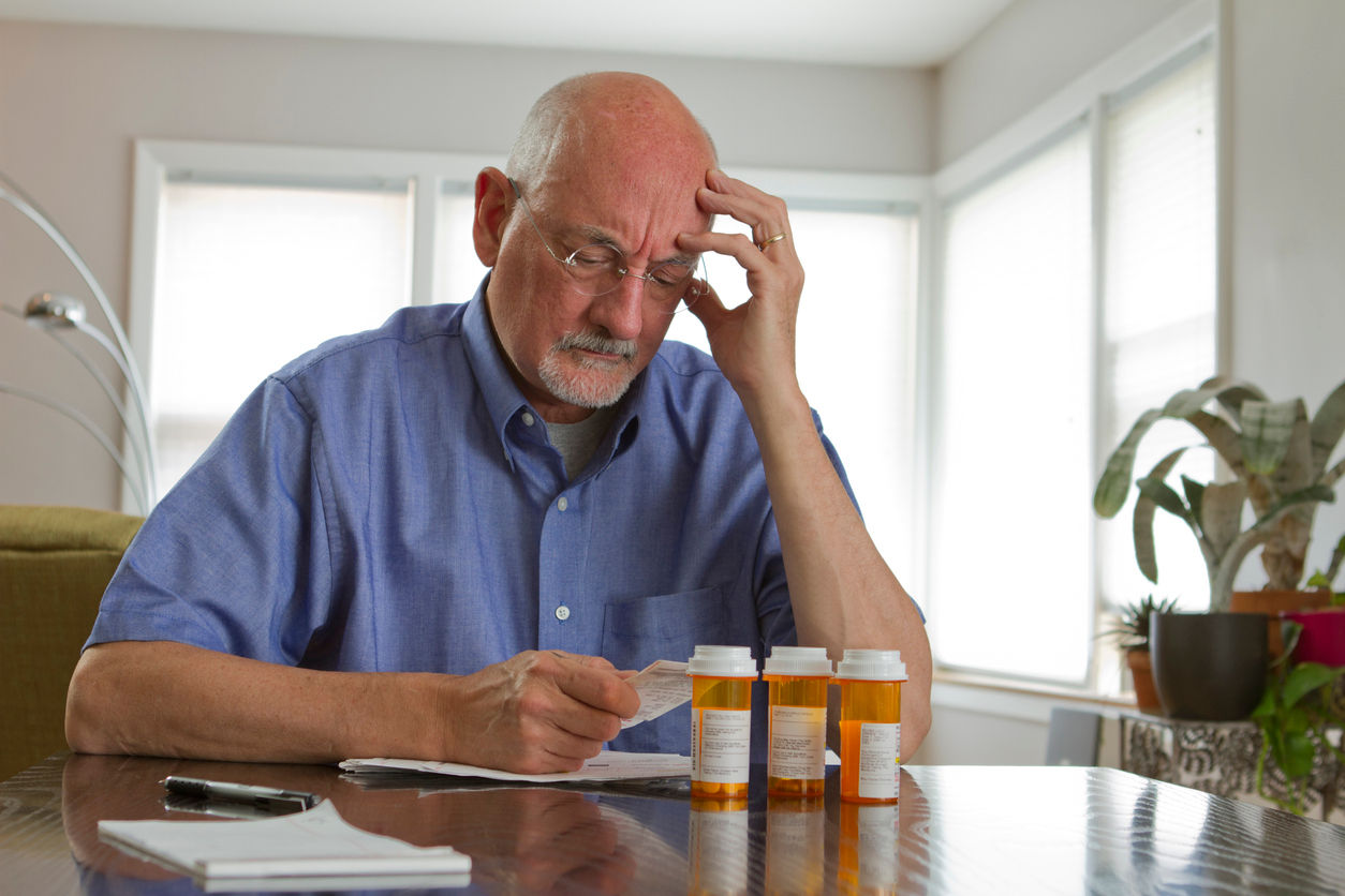 older man looking at bills and appearing stressed. next to him are prescription drug bottles