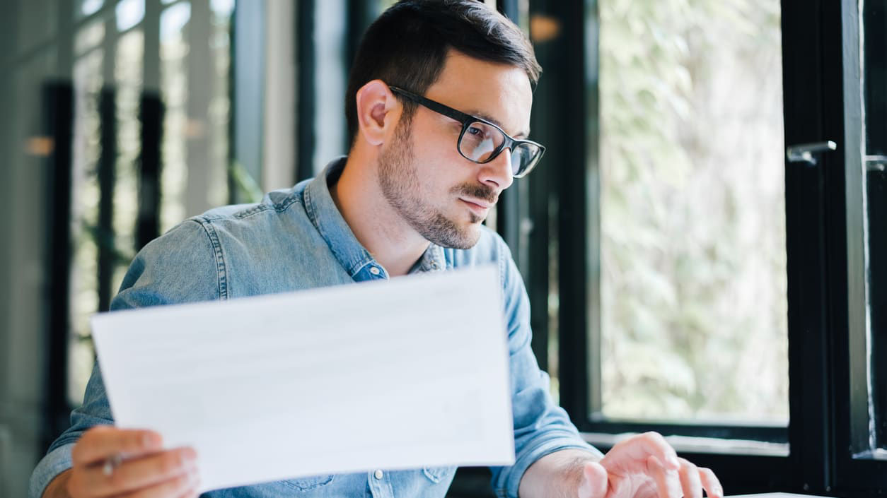 A man in glasses is looking at a piece of paper while sitting at a desk.