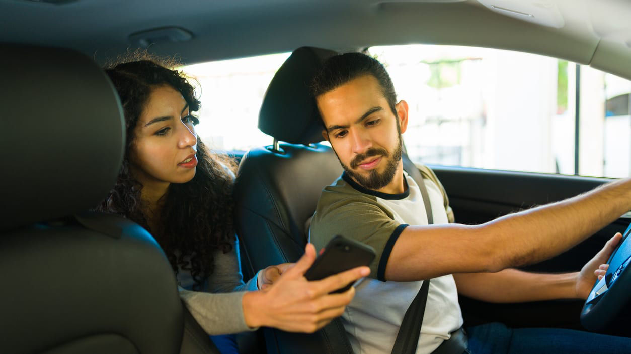 A man and woman in a car looking at their cell phone.