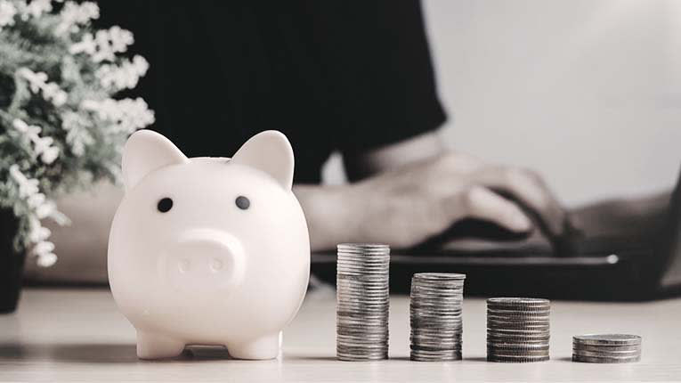 A piggy bank next to a pile of coins and a laptop.