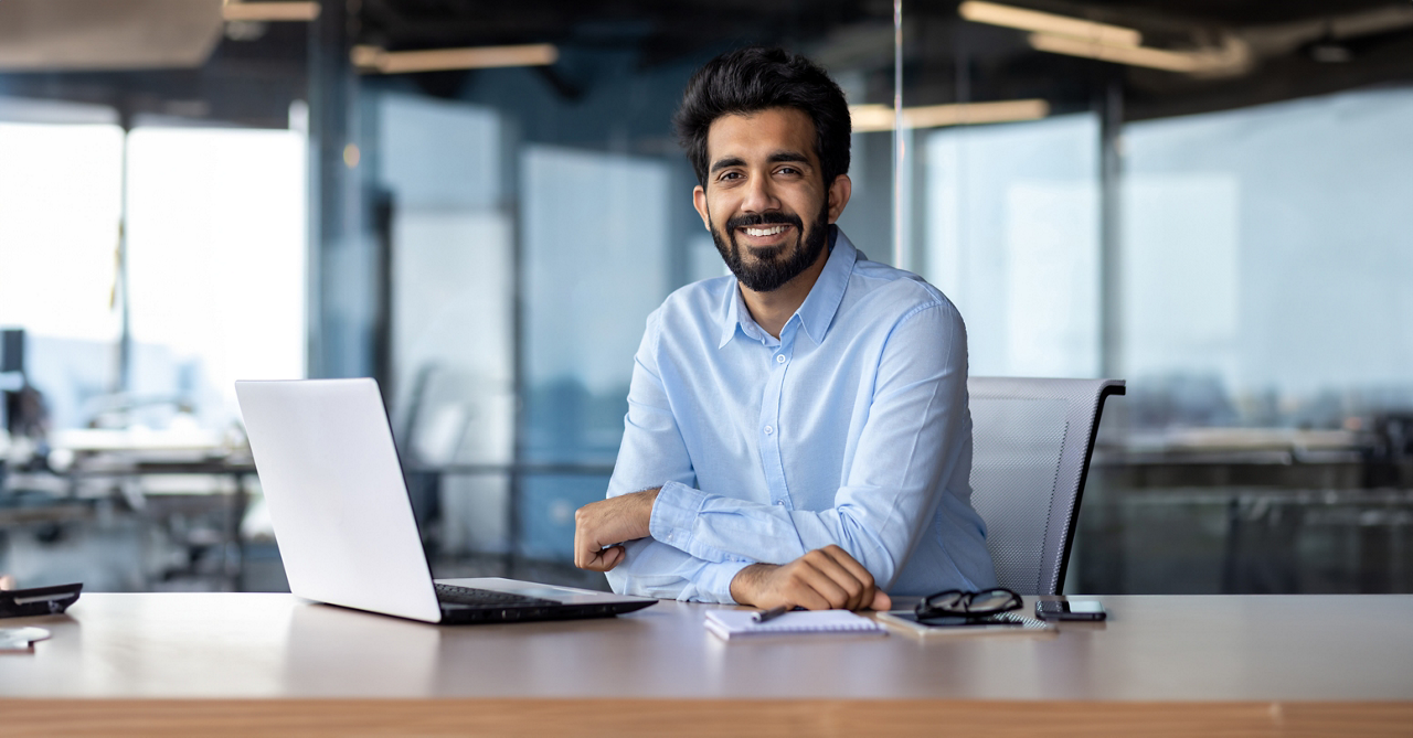 Portrait of happy and successful businessman, indian man smiling and looking at camera, satisfied with achievement results man working inside office building using laptop at work.
