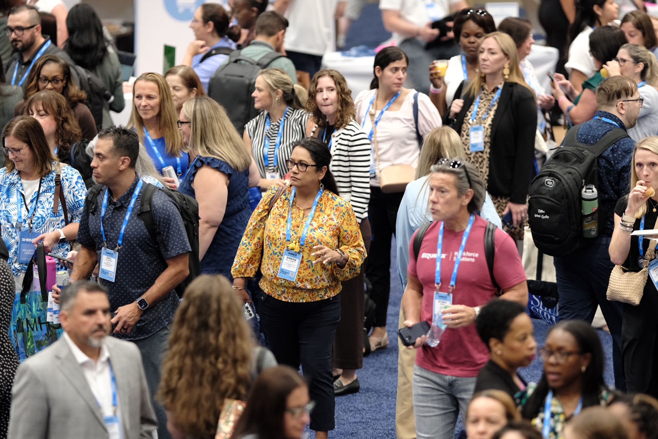 A crowd of attendees of SHRM26 walk through a packed hallway.. The attendees are wearing business casual clothing and engaging in thoughtful conversation.