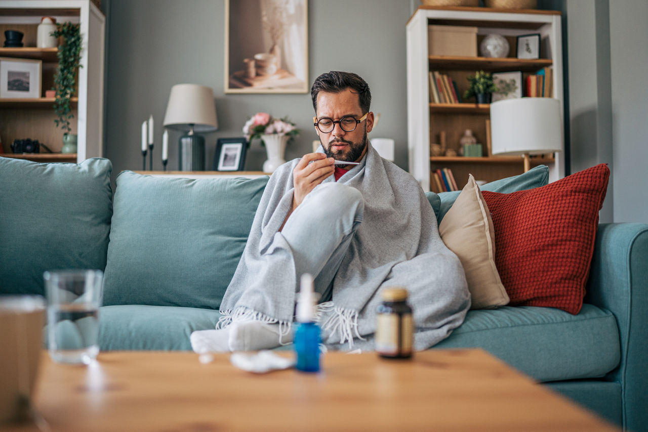 Sick man wrapped in blanket sitting on sofa in living room checking his temperature with thermometer, having flu symptoms, medicines on the table