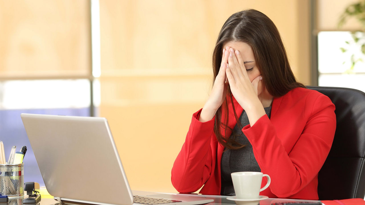 A woman is covering her face while sitting at a desk with a laptop.