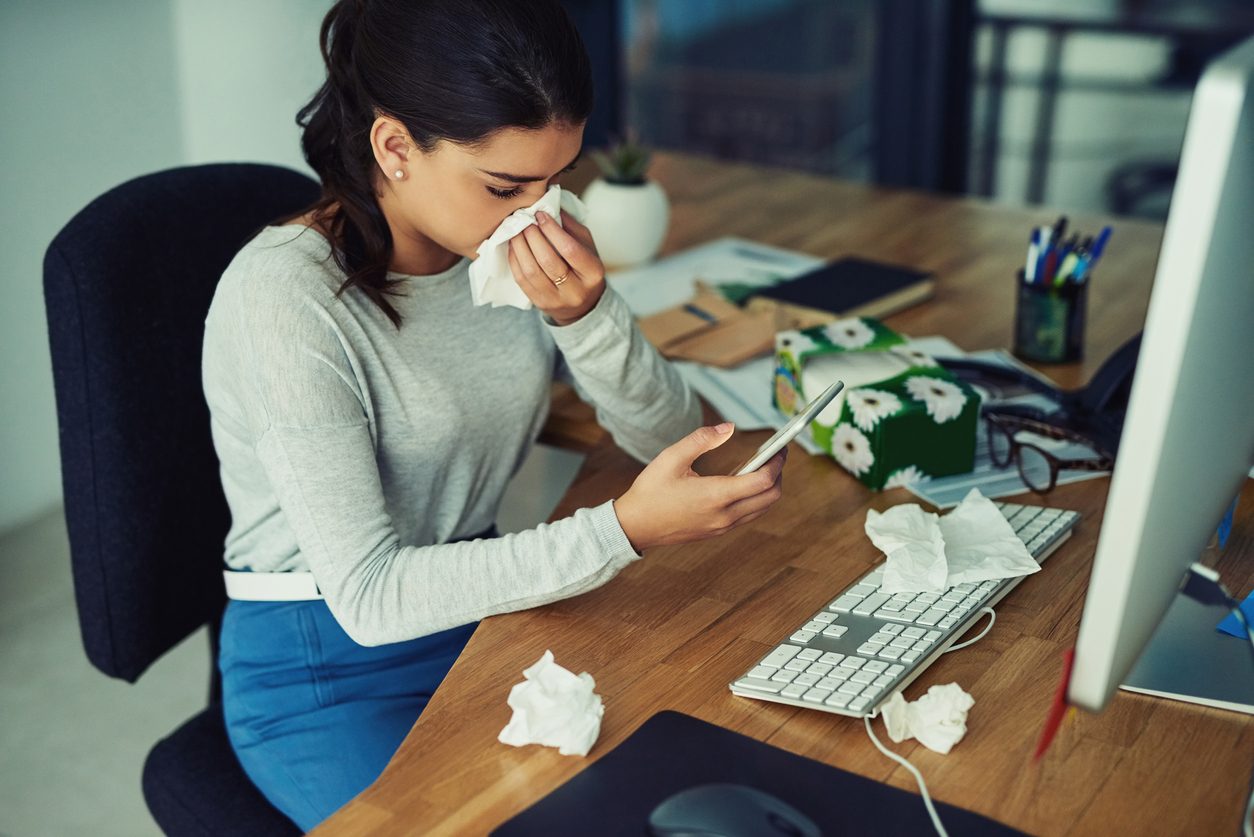 Shot of a businesswoman blowing her nose while looking at a cellphone in an office