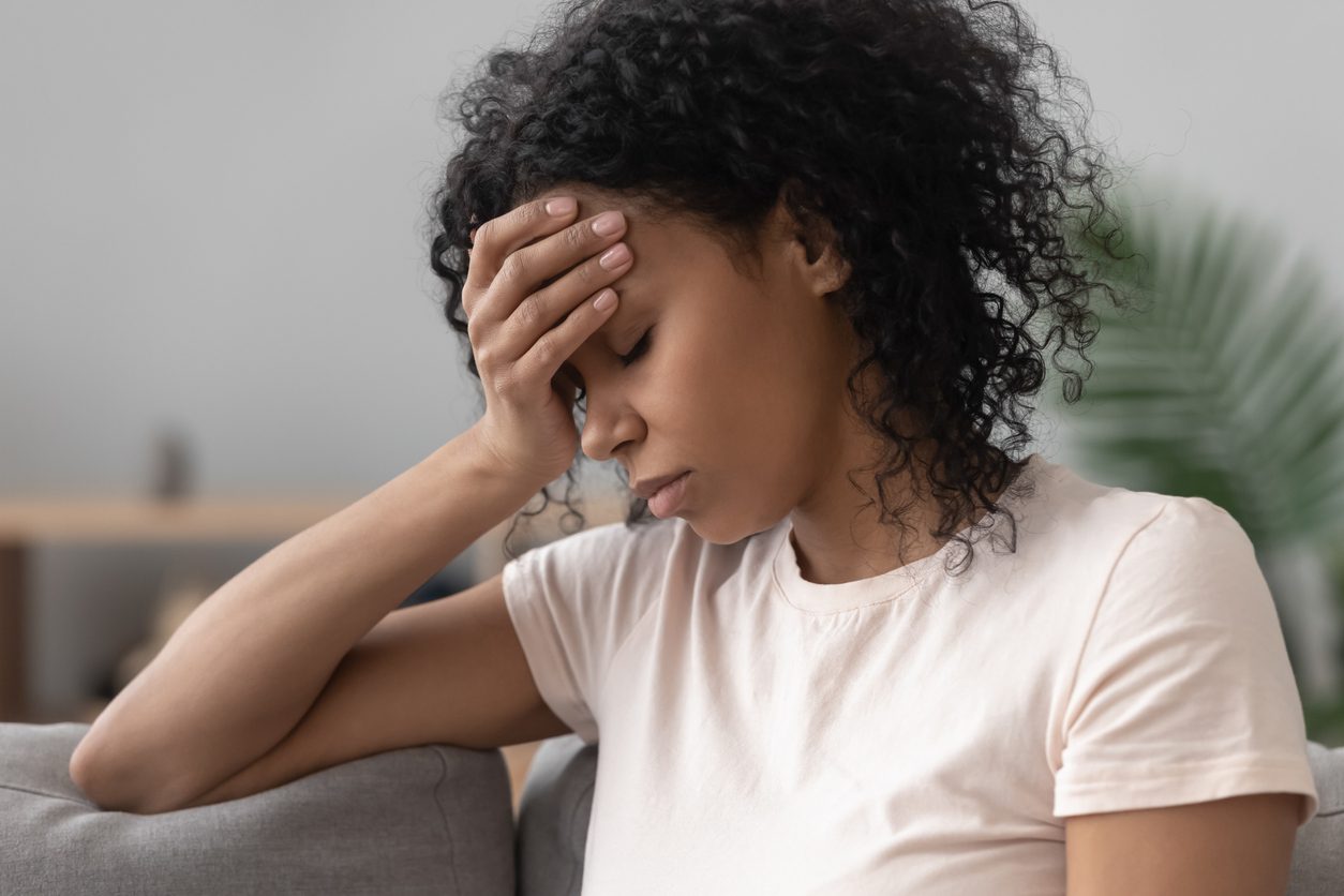 Close up African American woman sits on couch with hand to her forehead and eyes closed