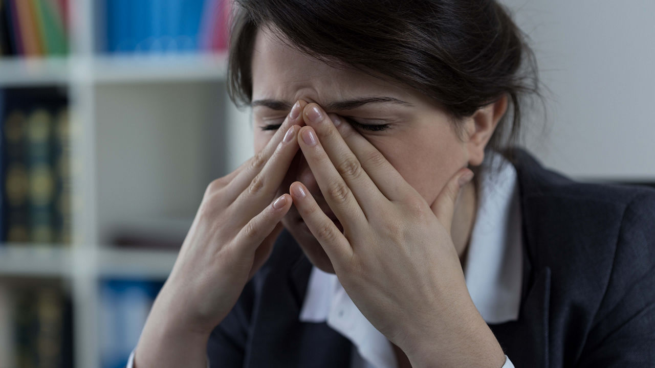A woman is covering her eyes while working on a laptop.