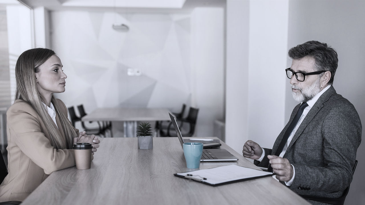 Women in suit on lefthand side sitting at table, man on right conducting interview and looking at resume