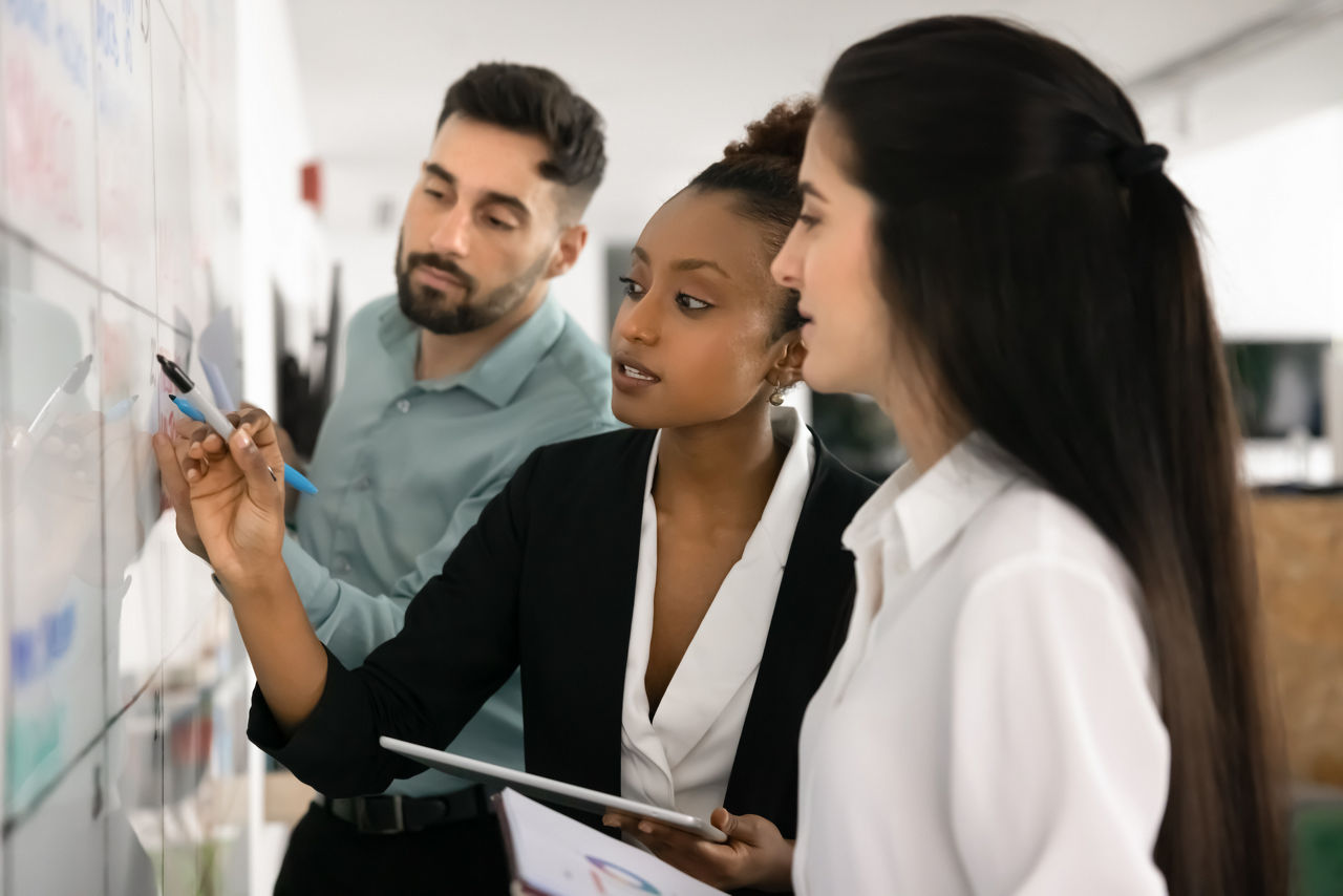 Diverse workmates working together on business plan, write on whiteboard standing in modern office, engaged in visual representation of data or market trends analysis.