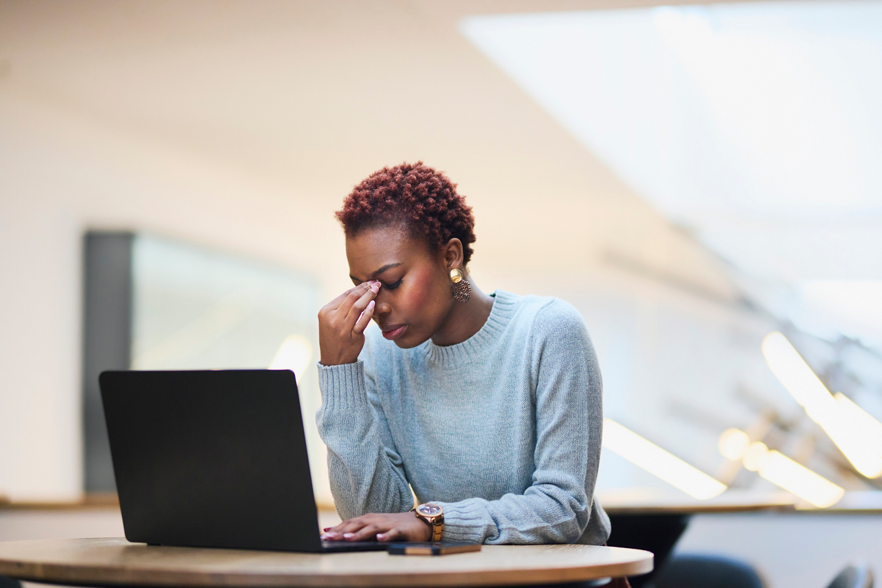 Close-up of a young Black woman in a light blue sweater, looking stressed and holding her forehead while working on a laptop at a round table indoors.