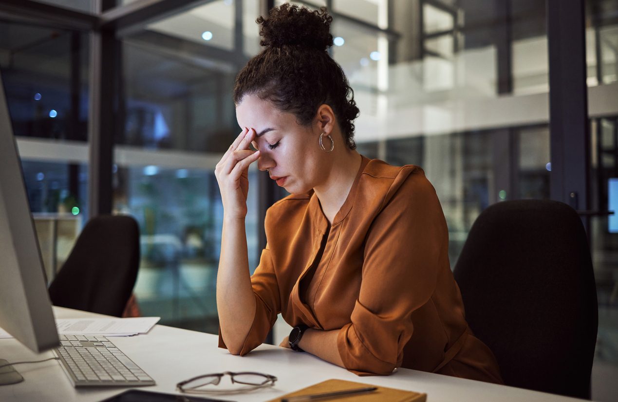 woman working at computer in office at night with fingers to forehead and eyes closed