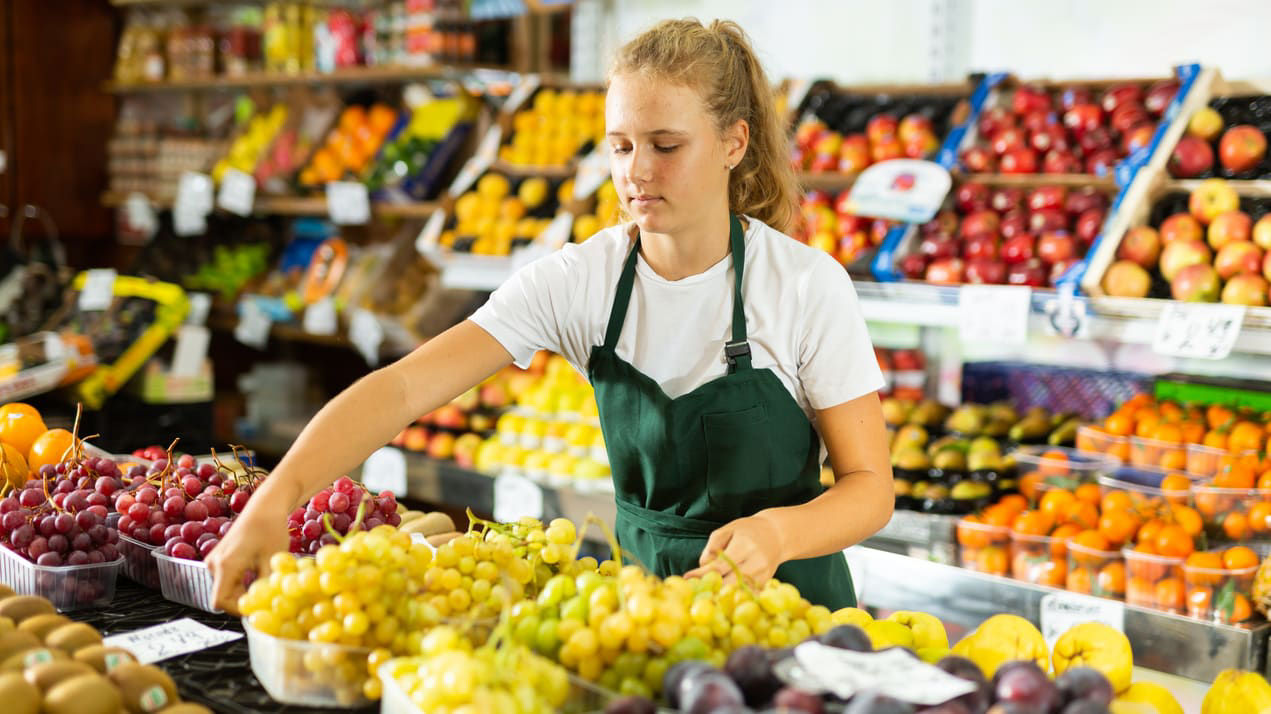 A woman in a green apron shopping for fruit in a grocery store.