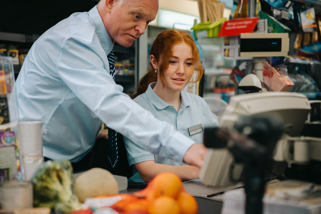Senior manager showing the checkout process to new female employee in grocery store. New cashier getting help from store manager at checkout counter.