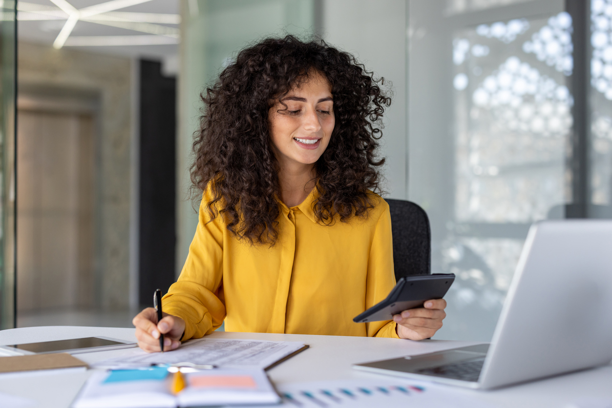 Latin American businesswoman accountant at office desk with calculator, pen, and laptop, demonstrating multitasking skills in financial work. Professional woman exemplifies confidence and efficiency.