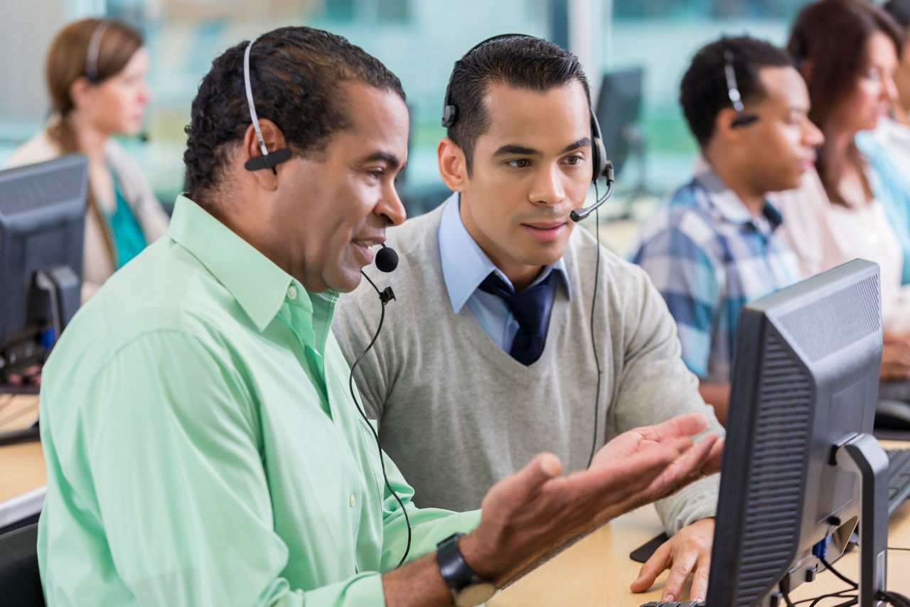 Helpful mature African American call center operator trains new male employee. The man gestures while showing the trainee something on his desktop computer. Both men are wearing headsets.