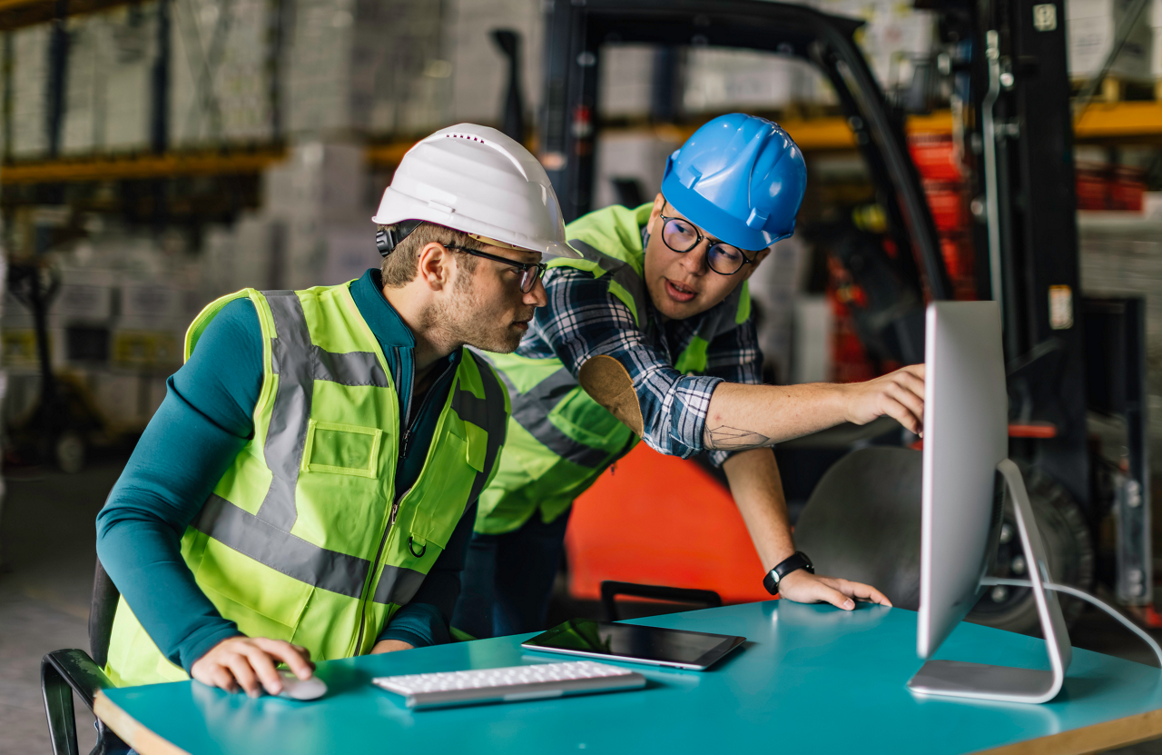 Two warehouse workers wearing safety helmets and high-visibility vests analyze data on a desktop computer in an industrial warehouse setting. One worker is pointing at the screen while the other listens attentively. In the background, a forklift and stacked inventory indicate a busy logistics and supply chain environment