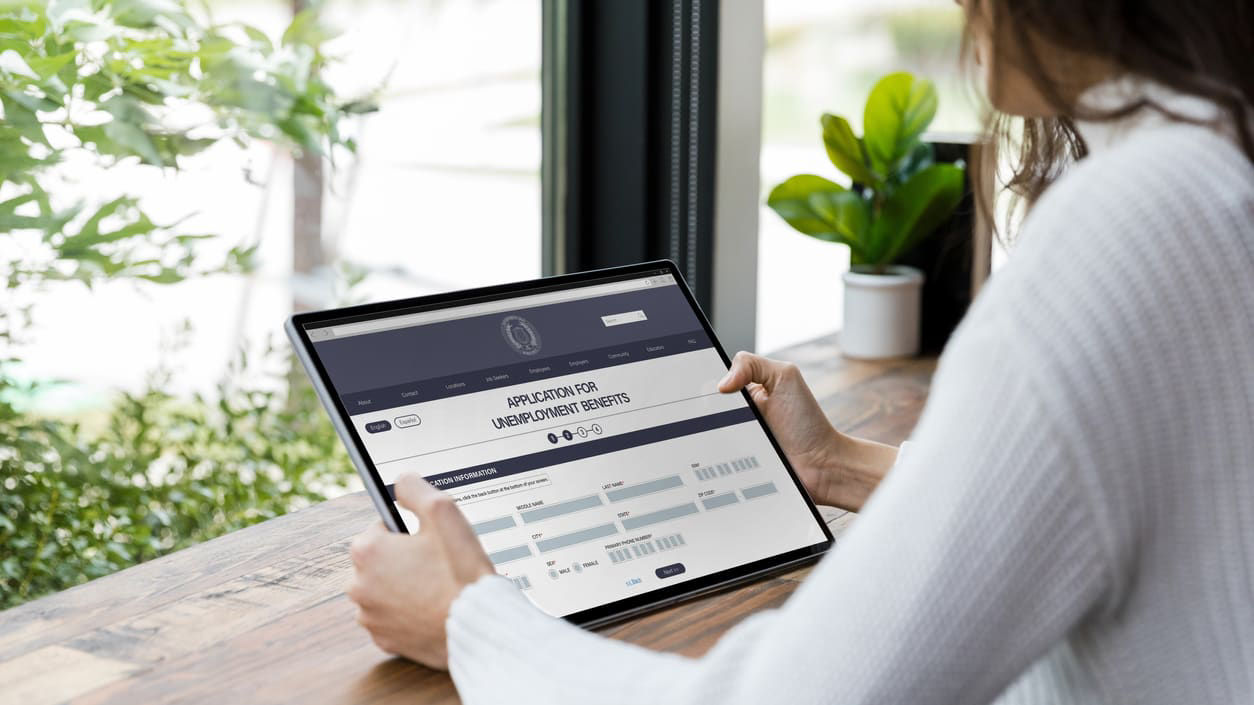 A woman is sitting at a table with a tablet computer in front of a window.