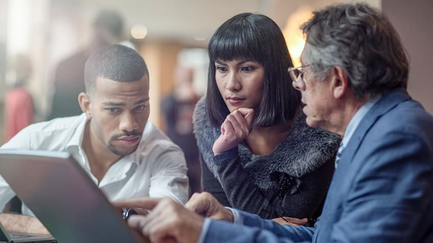Three people sitting around a table looking at a laptop.