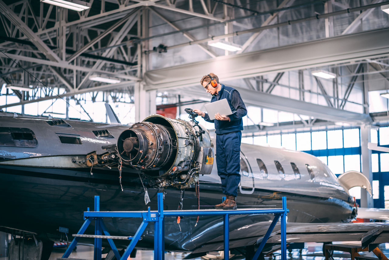 Aircraft mechanic standing on a raised platform, looking at a liquid crystal display of a probe inspection camera, trying to diagnose a fault in a jet engine of a small private airplane in an airport maintenance hangar.