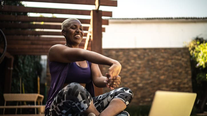 A woman is stretching on a yoga mat while using a laptop.