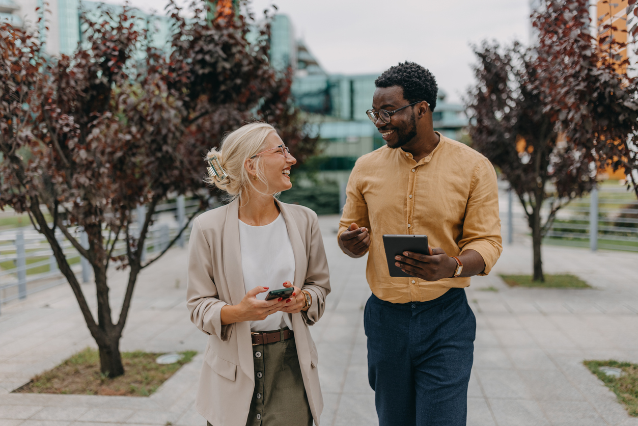 Image of two professionals walking in an urban area, engaged in a conversation while holding a smartphone and a tablet, indicative of a business meeting or collaboration.
